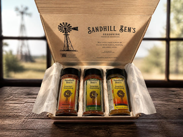 An open cardboard gift box from Sandhill Ben's Seasoning sits on a rustic wooden table in front of a blurred window. Inside the box are three spice jars with black lids and colorful labels, wrapped in white tissue paper.