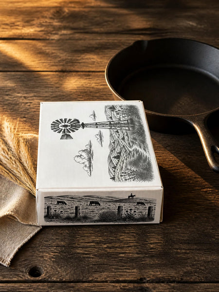 A white rectangular box with black rustic farm illustrations sits on a dark wooden table next to a black cast iron skillet and wheat stalks.