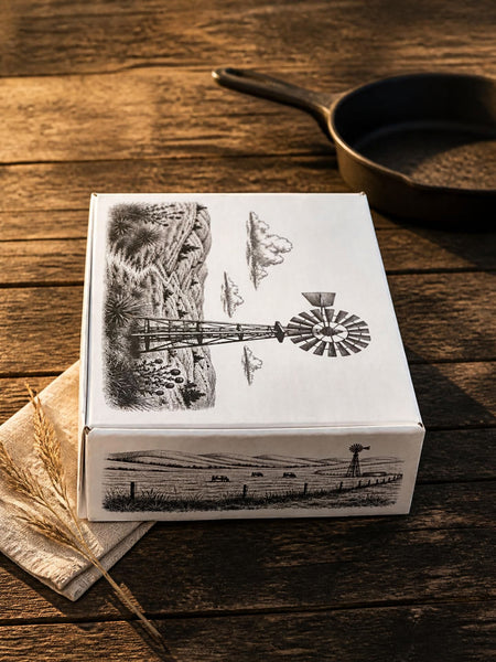 A white square product box with black rustic illustrations of a windmill, fence, and landscape, displayed on a dark wooden table next to a cast iron skillet and wheat stalks.